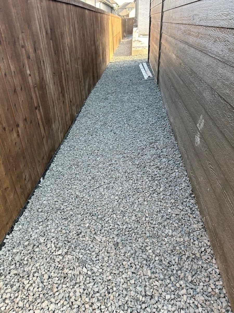 a narrow side yard: an open drainage trench and pipe, then the finished gravel walkway between two fences after