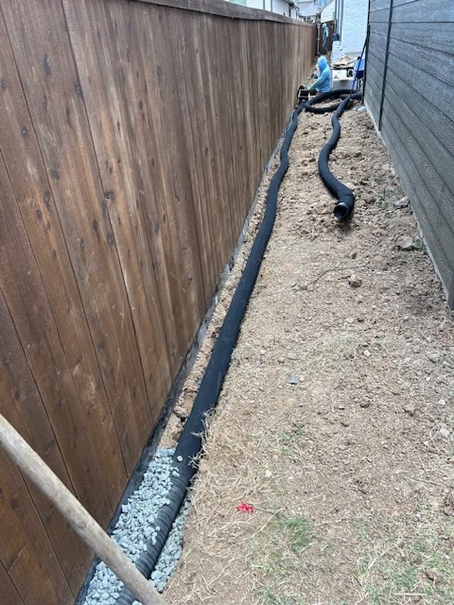 a narrow side yard: an open drainage trench and pipe, then the finished gravel walkway between two fences before